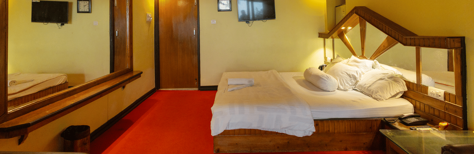 A bedroom with a king size bed, a wooden ceiling, a large mirror, a bedside table, and a wall mounted TV at Hotel Seagull Digha.